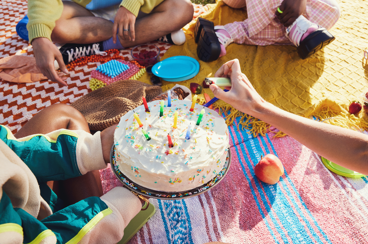 A group of friends sit around a cake; one friend is lighting birthday candles on the cake.